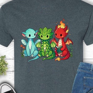 May include: A dark heather grey t-shirt with a graphic of three cartoon dragons. The dragons are green, blue, and red and are holding a book. The text "Enchanted Armory" is printed on the shirt.