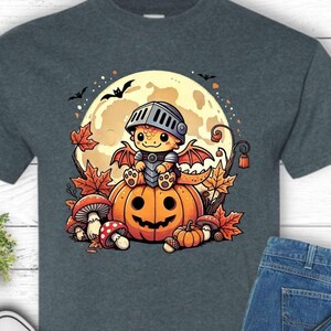 May include: Dark grey t-shirt with a Halloween graphic. The design shows a cartoon knight on a pumpkin, with a full moon and autumn leaves. The shirt is styled with blue jeans and black trainers. The image also includes the brand logo.