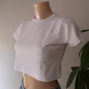 Könnte beinhalten: Ein schlichtes, weißes, kurzärmeliges bauchfreies Oberteil mit Rundhalsausschnitt. Das Kleidungsstück ist figurbetont geschnitten und hat einen geraden Saum, der über der Taille endet. Es wird über einer blauen Jeans mit sichtbaren Nähten getragen.