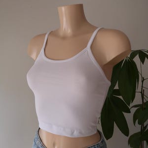 Op de afbeelding: Een effen wit kort hemdje met dunne schouderbandjes en een ronde halslijn, gedragen op een etalagepop-torso in combinatie met een lichtblauwe spijkerbroek. Het kledingstuk heeft een eenvoudig, aansluitend ontwerp met een rechte zoom.