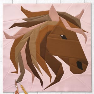 Könnte beinhalten: Ein geometrischer Pferdekopf-Quiltblock in Braun-, Beige- und Rosatönen. Mähne und Gesicht des Pferdes sind aus verschiedenen Stoffstücken gefertigt. Eine kleine, goldfarbene Schere und ein Werkzeug mit Holzgriff liegen neben dem Quilt.