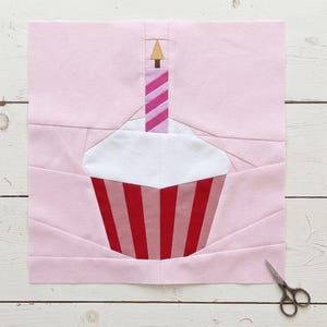 May include: A pink fabric block featuring a cupcake design with a lit candle. The cupcake has a white top, a red and pink striped base, and a pink and white striped candle with a gold flame. Small scissors are in the lower right corner.
