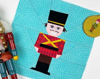 May include: A wooden nutcracker soldier with a red and blue uniform stands next to a teal fabric square with a nutcracker design. A small candle in a glass jar and a gold-colored ornament are also present. The scene evokes a festive holiday theme.