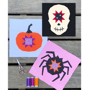 以下が含まれることがあります： ハロウィーンをテーマにした3つのファブリックブロック：カボチャ、スカル、クモ。カボチャはオレンジ色で、紫と青のスターバースト模様です。スカルは白で、赤と黒のスターバースト模様です。クモは黒で、オレンジと紫のスターバースト模様です。ハサミと糸巻きも含まれています。
