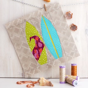 May include: A beige fabric wall hanging with two colorful surfboards, one green and one blue, with a white background. The surfboards are made of fabric and have a patchwork design. The wall hanging is hanging from a white string with a wooden clothespin. There are seashells on the white wooden background.