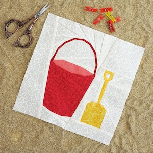 May include: A white fabric square with a red bucket and yellow shovel design. The fabric has a white heart pattern. The square is on a brown sandy surface with red and yellow clothespins scattered around it.