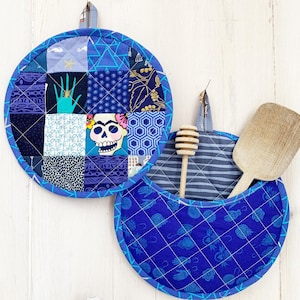 Könnte beinhalten: Zwei gesteppte blaue Topflappen mit Aufhängeschlaufen. Einer hat ein Patchwork-Design mit einem von Frida Kahlo inspirierten Totenkopf, der andere hat eine Tasche mit einem Holzlöffel und einem Honiglöffel. Drei Garnrollen in Creme und Blau im Vordergrund.