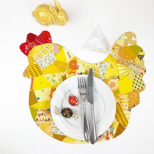 Puede incluir: Un mantel individual amarillo con forma de gallina, cresta roja y diseño patchwork. Un plato blanco con cubiertos y caramelos envueltos se encuentra sobre el mantel. Una figura de conejo dorado y una copa transparente completan la mesa festiva.