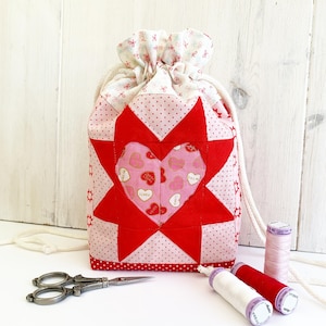 May include: A handmade drawstring bag with a red star and heart design. The bag is made of pink and white patterned fabric, with a white drawstring closure. A pair of scissors and spools of thread are next to the bag.