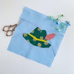 Pode incluir: Um quadrado de tecido azul com bolinhas brancas e uma aplicação de tecido verde, amarelo e vermelho de um chapéu com uma pena. O quadrado de tecido está preso com clipes de plástico transparentes.