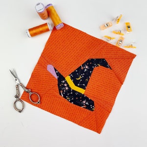 Könnte beinhalten: Ein Hexenhut-Patchworkblock mit einem schwarzen, gelben und rosafarbenen Stoffdesign. Der Block ist von orangefarbenem Stoff mit einem sich wiederholenden Textmuster umgeben. Es gibt auch Spulen mit orangefarbenem Garn und orangefarbene Nähklammern.