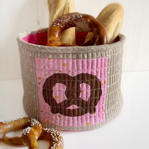 Op de afbeelding: Een roze en bruine quilted stoffen mand met een pretzel-ontwerp. De mand is gevuld met brood en pretzels.