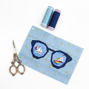May include: A light blue fabric patch featuring sunglasses with sailboat details. Two spools of thread in light blue and navy blue are positioned above the patch. A pair of silver scissors is placed to the left of the patch.
