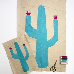 May include: Two fabric cactus quilt blocks, one large and one small, in turquoise and pink on a tan background. The large block is approximately 50 cm tall. Sewing supplies, including scissors and thread spools, are also present.