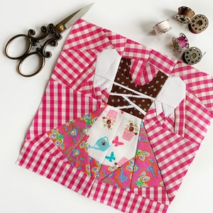 Peut inclure: Un carré de tissu vichy rose et blanc avec un motif de patchwork représentant une robe, des fleurs et des papillons. Le tissu est entouré d'accessoires de couture, notamment des ciseaux, des bobines de fil et une aiguille.