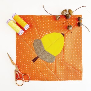 May include: Orange fabric with a patchwork design of an acorn. The fabric is decorated with small white dots. Two spools of yellow thread are in the top left corner of the image.