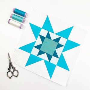Puede incluir: Un bloque de edredón de estrella blanco y azul con un diseño de estrella turquesa. También hay tres carretes de hilo en tonos azules y un par de tijeras pequeñas. El bloque de edredón tiene forma cuadrada.