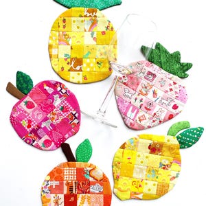 Puede incluir: Un conjunto de posavasos con forma de fruta y acolchados. El juego incluye una piña, una fresa, una manzana y un limón, cada uno hecho de cuadrados de patchwork en varios tonos de amarillo, rosa, naranja y verde. Una copa de vino descansa entre los posavasos.