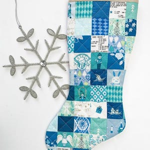 May include: A quilted Christmas stocking featuring a patchwork design of blue, teal, and white squares with holiday motifs like snowmen, trees, and cake recipes, paired with a silver wire snowflake ornament with a central clear gem.
