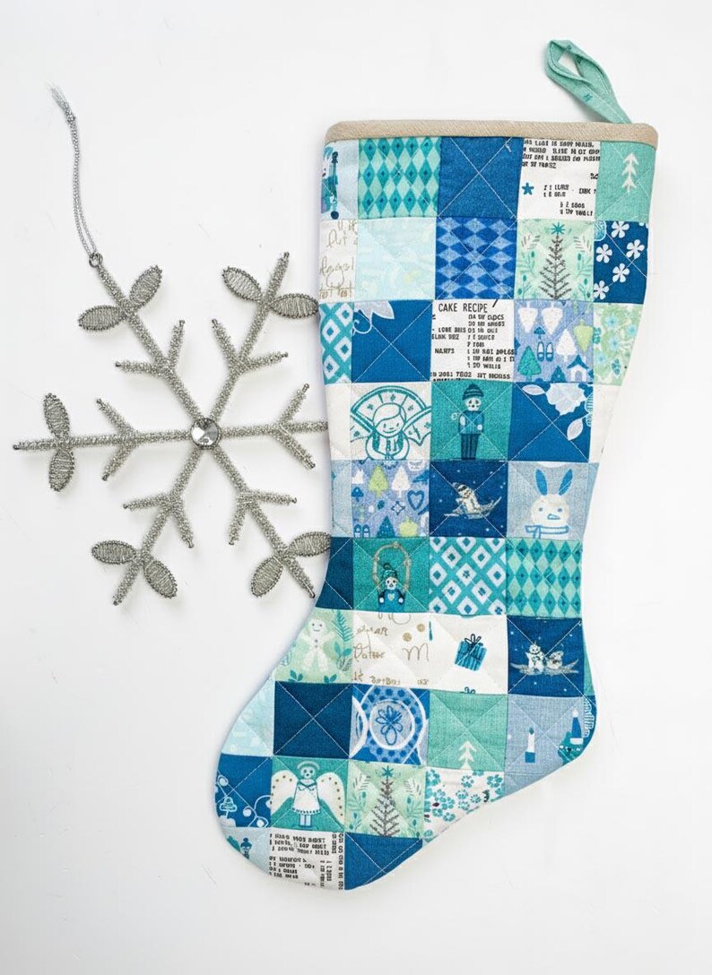 May include: A quilted Christmas stocking featuring a patchwork design of blue, teal, and white squares with holiday motifs like snowmen, trees, and cake recipes, paired with a silver wire snowflake ornament with a central clear gem.