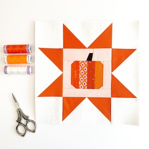 May include: A white fabric square with an orange star design. The star has a smaller pink square in the centre with a patchwork pumpkin design. There are three spools of orange thread and a pair of silver scissors on a white surface.