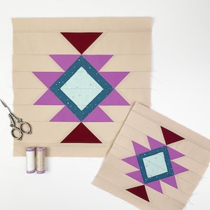 May include: Two beige fabric quilt blocks with a starburst design. The starburst features purple, teal, and burgundy geometric shapes. A pair of scissors and two spools of thread are also present.