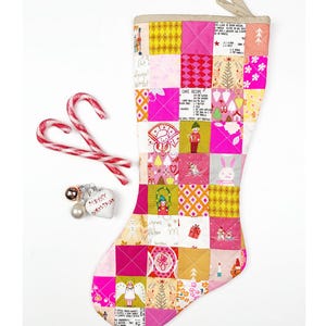 May include: A patchwork Christmas stocking in pink, yellow, and white, with festive images and text, including a cake recipe. Two candy canes and a "Merry Christmas" ornament are next to the stocking. The stocking is a festive holiday decoration.