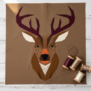 Può includere: Un blocco di quilt a forma di testa di cervo geometrica nei toni del marrone, beige e bordeaux. Il cervo presenta un design patchwork con vari modelli e trame. Le bobine di filo in colori coordinati sono posizionate vicino al blocco di quilt, suggerendo un progetto di cucito.