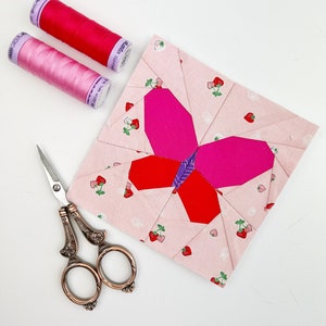 Könnte beinhalten: Ein rosa Stoffquadrat mit einem rosa und roten Schmetterlingsdesign und einem weißen Hintergrund mit kleinen Erdbeer-Drucken. Das Stoffquadrat ist von zwei Spulen mit rosa und rotem Garn und einer antiken Schere umgeben.