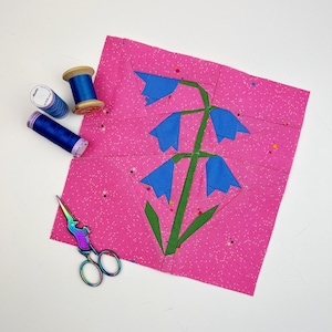 May include: A pink fabric square with a blue flower design. The flower has a green stem and five blue petals. The fabric has a white polka dot pattern.