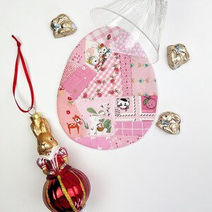 May include: A red and gold bunny ornament with a red ribbon hangs next to a pink patchwork egg-shaped coaster. A clear glass sits atop the coaster, and several foil-wrapped chocolate bunnies are scattered around the scene. The items are arranged on a white surface.