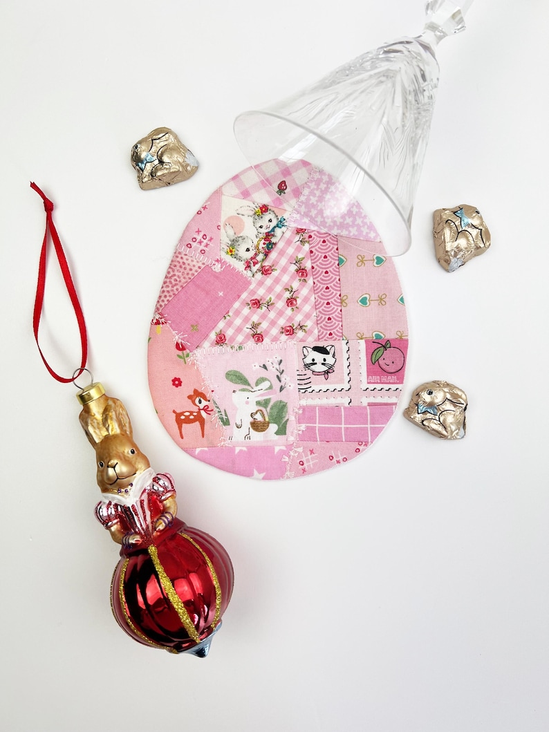 May include: A red and gold bunny ornament with a red ribbon hangs next to a pink patchwork egg-shaped coaster. A clear glass sits atop the coaster, and several foil-wrapped chocolate bunnies are scattered around the scene. The items are arranged on a white surface.