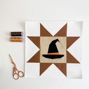 May include: A square quilt block featuring a central beige square with a black witch hat appliqué, surrounded by brown and white star points. Two spools of thread in dark brown and gold, and a pair of copper-colored scissors are placed nearby.