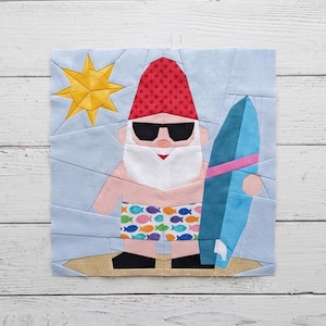 May include: A fabric quilt block featuring a gnome wearing a red star-patterned hat, black sunglasses, and swim trunks with a colorful fish print. The gnome holds a blue surfboard on a sandy beach under a yellow sun against a light blue sky.