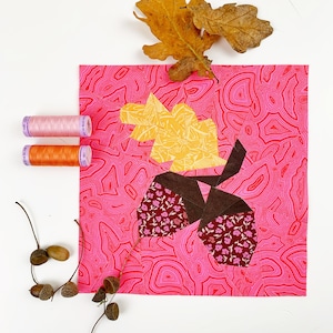 May include: A pink fabric square with a brown, pink, and yellow acorn design. There are three spools of thread, two orange and one pink, and several acorns on the white background.