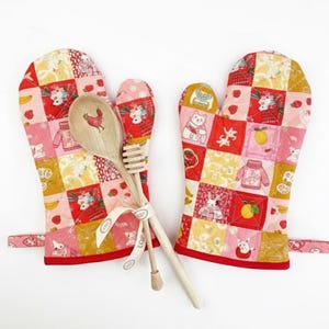May include: A pair of quilted oven mitts with a matching wooden spoon and honey dipper. The mitts are red, pink, and yellow with a patchwork design featuring strawberries, cats, and other whimsical illustrations. The spoon has a rooster design.