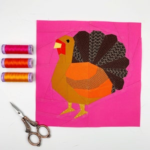 Op de afbeelding: Een roze stoffen achtergrond met een kleurrijke patchwork-kalkoen-applique. De kalkoen is gemaakt van bruin, oranje, geel en rood stof. De kalkoen heeft een bruine staart met gouden accenten, een rode kop en gele poten.
