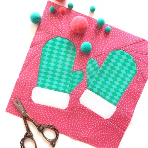 Puede incluir: Un cuadrado de tela rosa con un patrón de lunares blancos. Dos mitones verdes y blancos están aplicados en la tela. La tela está rodeada de pompones rosas y verdes. Un par de tijeras antiguas está sobre la tela.