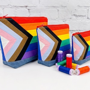 Peut inclure: Trois pochettes zippées de tailles décroissantes, chacune arborant un motif de drapeau en chevron avec des bandes rouges, orange, jaunes, vertes, bleues, violettes, marron, noires, blanches, roses et bleu clair, sur une base en jean bleu.