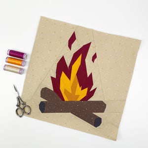 Könnte beinhalten: Ein Stoffblock mit einem Lagerfeuer-Design. Die Flammen bestehen aus rotem und gelbem Stoff, mit braunen Baumstämmen an der Basis. Der Hintergrund ist ein beigefarbener Stoff mit kleinen weißen und goldenen Punkten. Drei Garnrollen und eine Schere sind ebenfalls sichtbar.