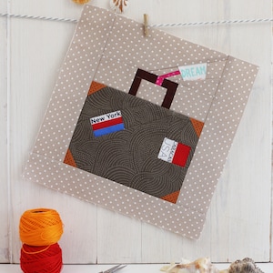May include: A beige fabric square with white polka dots, featuring a brown suitcase with a red, white and blue tag that says "New York" and a red tag that says "Caracas, USA". The suitcase has a brown handle and a white tag that says "Dream".