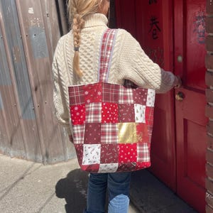 May include: A patchwork tote bag featuring a variety of red, white, and gold fabric squares with holiday-themed patterns including snowflakes, candy canes, and plaid designs, equipped with two matching fabric shoulder straps.