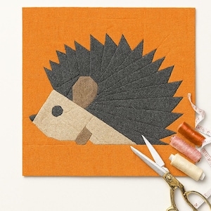 Könnte beinhalten: Ein handgefertigtes Igel-Kunstwerk auf orangefarbenem Hintergrund. Der Igel besteht aus Stoff in Schwarz-, Beige- und Brauntönen. Goldene Schere, Garnrollen und ein Maßband sind um das Kunstwerk angeordnet, was auf ein Bastelprojekt hindeutet. Maße in cm.