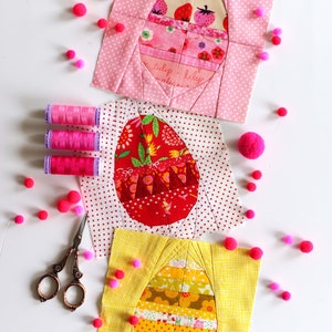 May include: Three fabric patchwork squares with a pink, red, and yellow background. Each square features a different design of an egg with a variety of colorful patterns. The squares are surrounded by pink pom-poms.