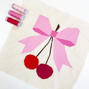 May include: A pink fabric bow with two red and pink cherries on a white background with pink polka dots.