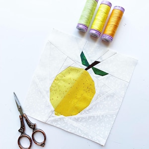 Puede incluir: Un bloque de tela hecho a mano con un limón amarillo y hojas verdes, junto con tres carretes de hilo en tonos amarillos y verdes. Unas tijeras ornamentadas descansan cerca. El bloque está sobre un fondo blanco.