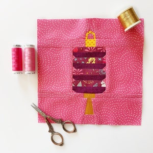 Könnte beinhalten: Ein rosa Stoffquadrat mit einem Patchwork-Design einer Laterne. Die Laterne besteht aus violettem, gelbem und floralem Stoff. Das Stoffquadrat ist von Garnrollen und einer Schere umgeben.