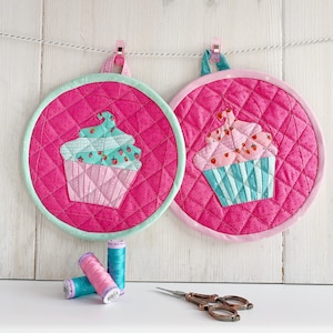 Może przedstawiać: Dwa różowe pikowane uchwyty na gorące z wzorem zielonego i różowego ciastka. Uchwyty są zawieszone na białym sznurku z różowymi klipsami. Na pierwszym planie znajdują się trzy szpulki nici.