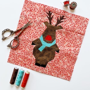 Może przedstawiać: Aplikacja renifera w kolorze brązowym i czerwonym z niebieskim szalikiem na różowo-białym materiale w kwiaty. Materiał otoczony jest przyborami do szycia, w tym szpulkami nici i nożyczkami.