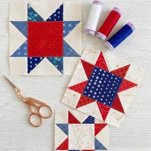 May include: Three completed star quilt blocks in red, white, and blue fabrics. The blocks are arranged with spools of white, red, and blue thread and a pair of gold scissors. The blocks are on a white wooden surface.
